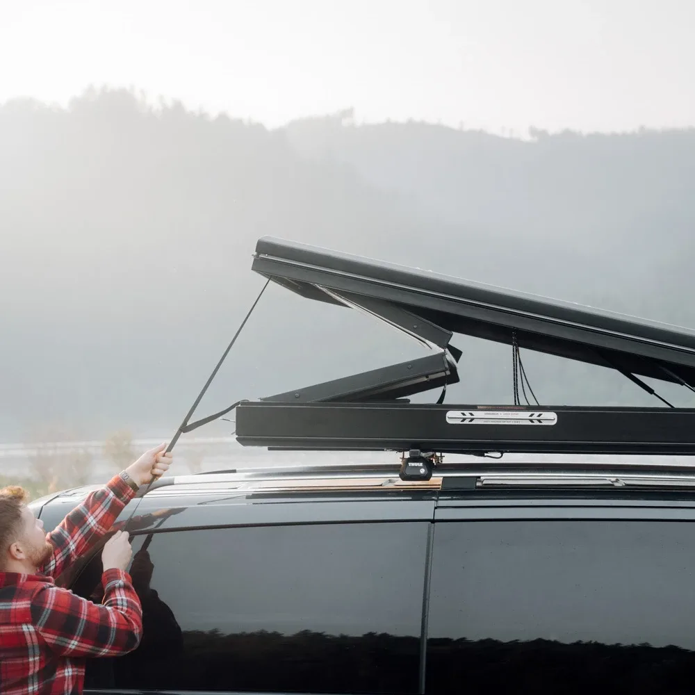 Person beim Öffnen eines Hardside-Dachzelts Dark Barracks Grand auf einem schwarzen Van vor nebliger Bergkulisse.