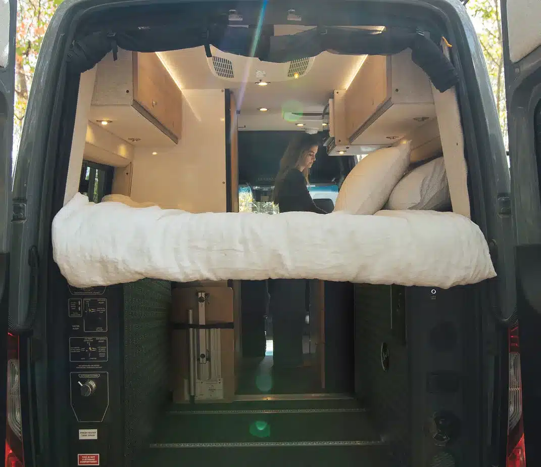 Dark MODE XO Kastenwagen mit hochgeklapptem Bett, Innenansicht durch geöffnete Hecktüren, Frau im Innenraum.