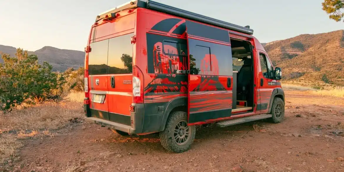 Roter RAM Promaster 2500 High Roof Kastenwagen mit auffälliger Grafikbeklebung, geöffneter Schiebetür und Campingausstattung in einer landschaftlichen Umgebung