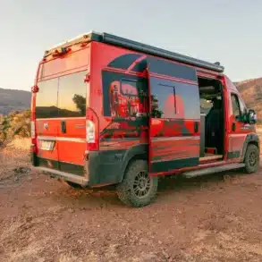 Roter RAM Promaster 2500 High Roof Kastenwagen mit auffälliger Grafikbeklebung, geöffneter Schiebetür und Campingausstattung in einer landschaftlichen Umgebung