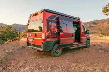 Roter RAM Promaster 2500 High Roof Kastenwagen mit auffälliger Grafikbeklebung, geöffneter Schiebetür und Campingausstattung in einer landschaftlichen Umgebung