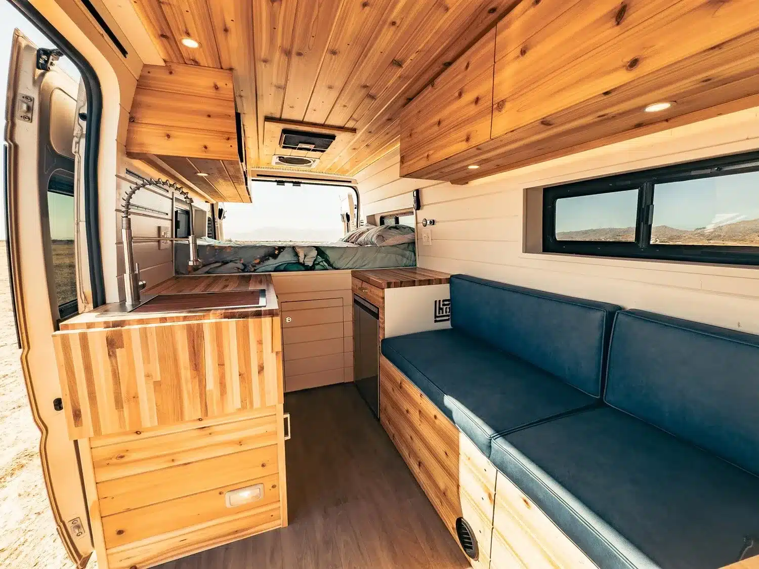 Innenansicht des Faith RAM Promaster Camper Van mit Holzverkleidung, blauer Sofa-Zone, Küchenzeile und Bett im Hintergund.