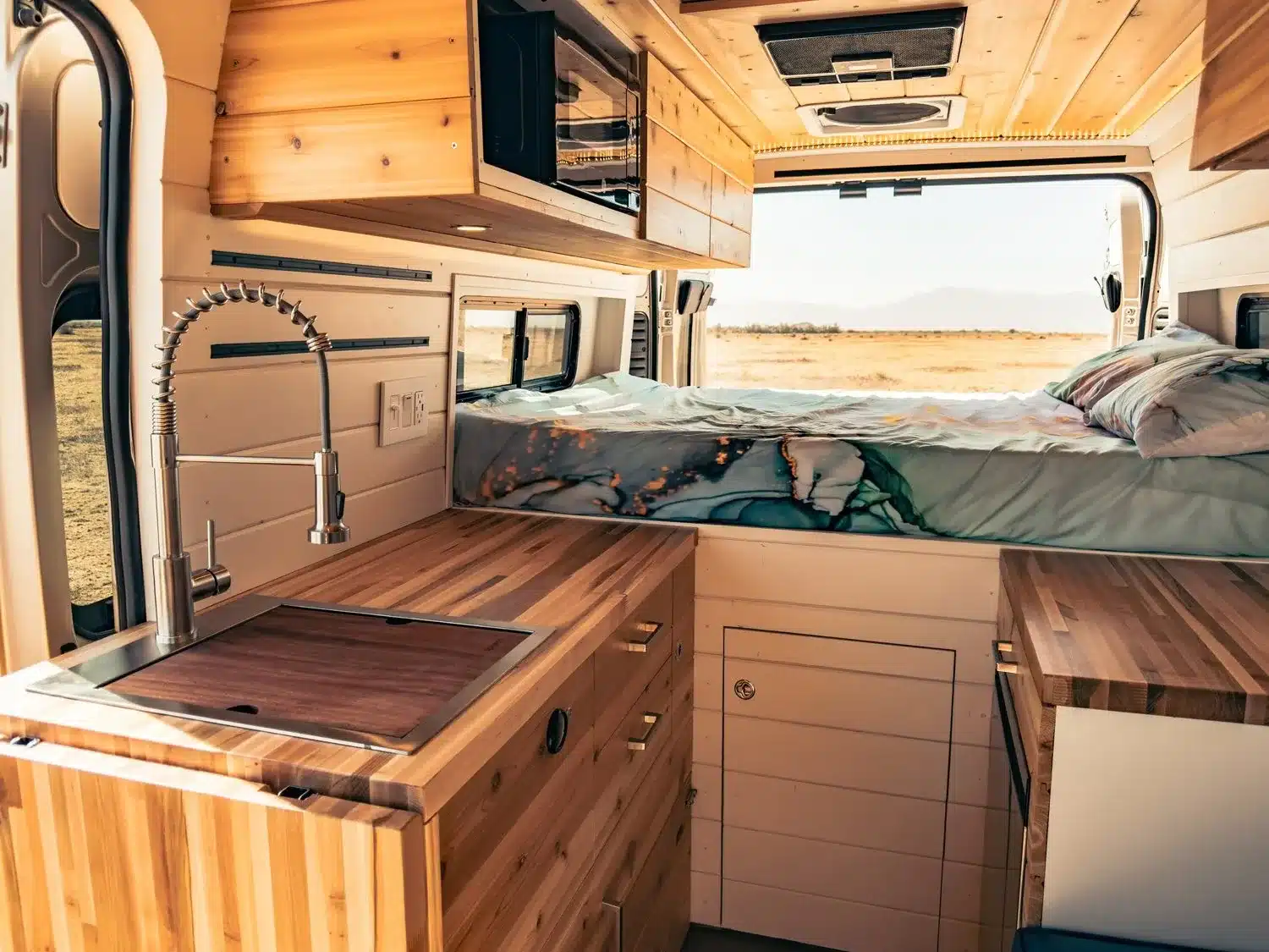 Küche und Schlafbereich im Faith RAM Promaster Camper Van mit Holzoberflächen, großem Fenster und Bett
