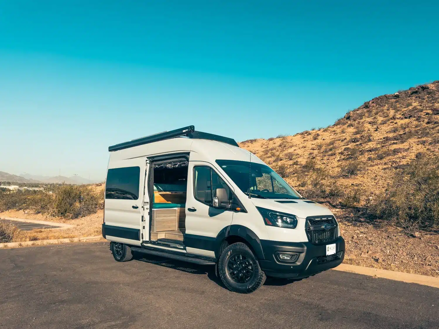 Ford Transit Trail Kastenwagen-Camper MJÖLNIR mit geöffnetem Seiteneinstieg, Hochdach und Dachträger auf einem Parkplatz vor Naturkulisse