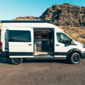 Seitenansicht eines weißen Ford Transit Trail 350 Campervans mit geöffneten Schiebetüren und sichtbarem Innenausbau vor Felskulisse