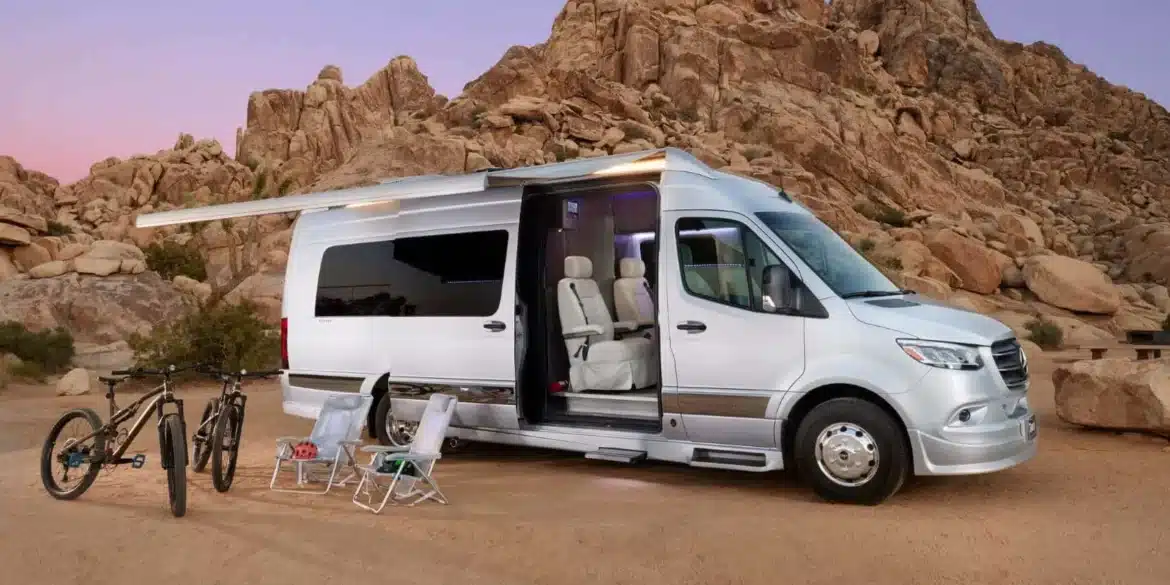 Silberner Grech STRADA-ion AWD Luxus-Campervan mit ausgefahrener Markise, Campingmöbeln und Fahrrädern vor Felsen