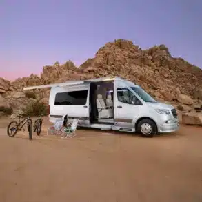 Silberner Grech STRADA-ion AWD Luxus-Campervan mit ausgefahrener Markise, Campingmöbeln und Fahrrädern vor Felsen