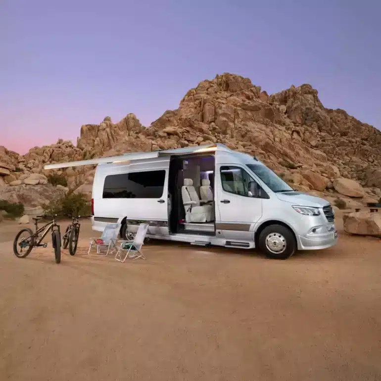 Silberner Grech STRADA-ion AWD Luxus-Campervan mit ausgefahrener Markise, Campingmöbeln und Fahrrädern vor Felsen