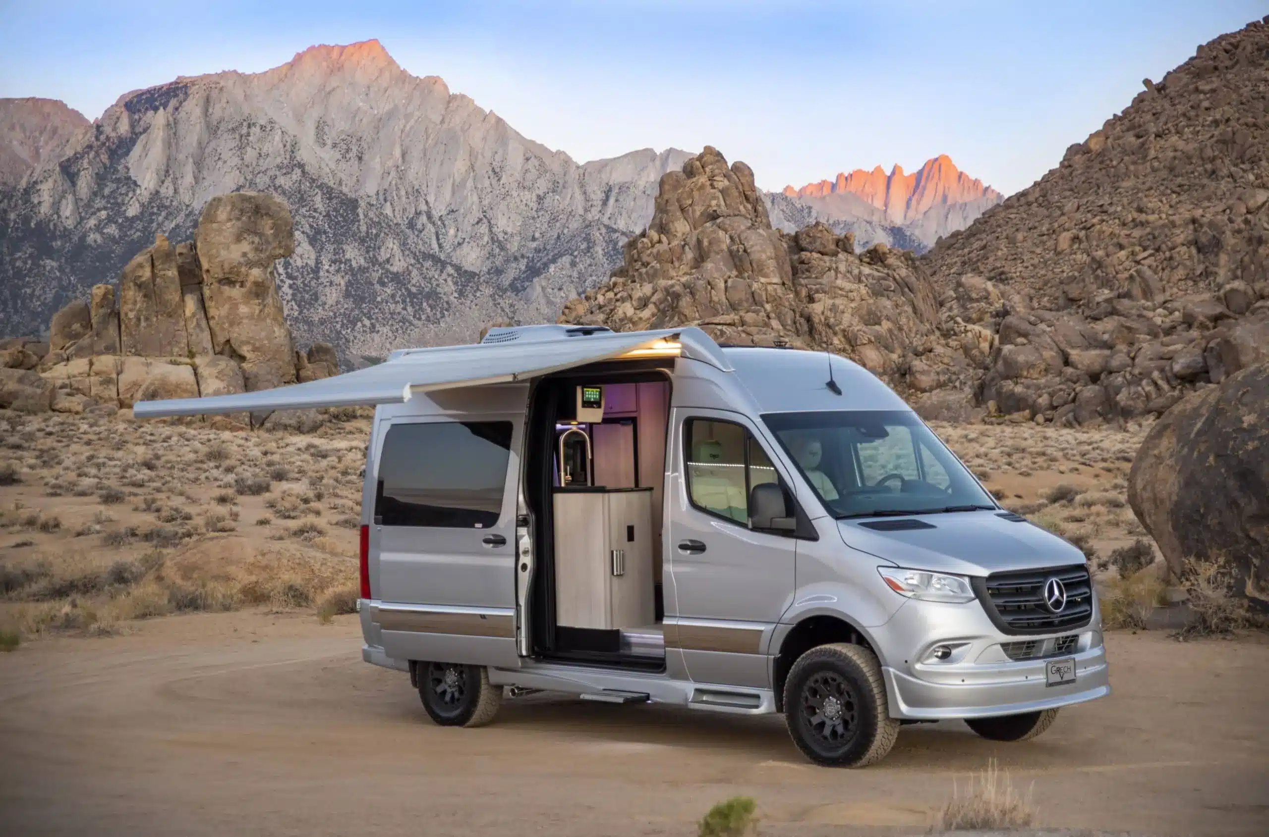 Silberner Grech Turismo-ion AWD 2026 Campervan auf Mercedes-Benz Sprinter-Basis mit geöffneter Seitentür und Markise vor felsiger Bergkulisse.