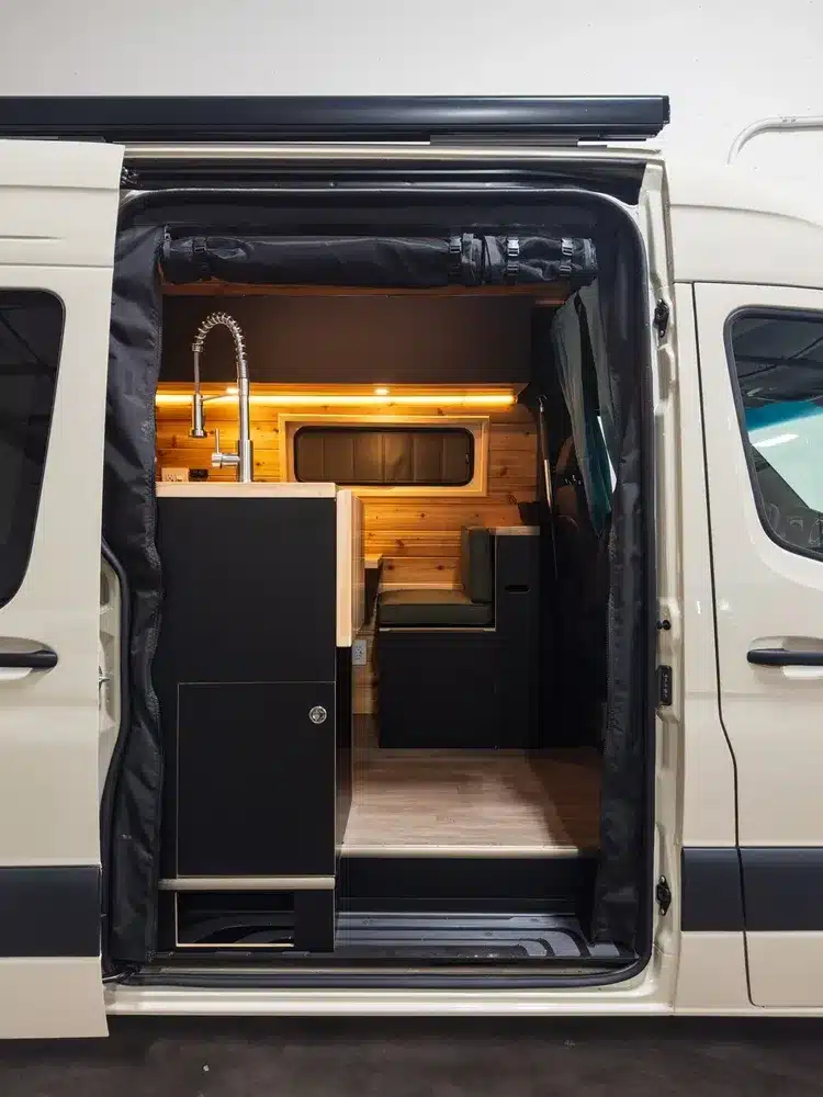 Seitlicher Eingang des Hive Hideaway Campervans zeigt Küchenblock mit Spüle und moderne Sitzgruppe aus Holz und schwarzen Elementen.
