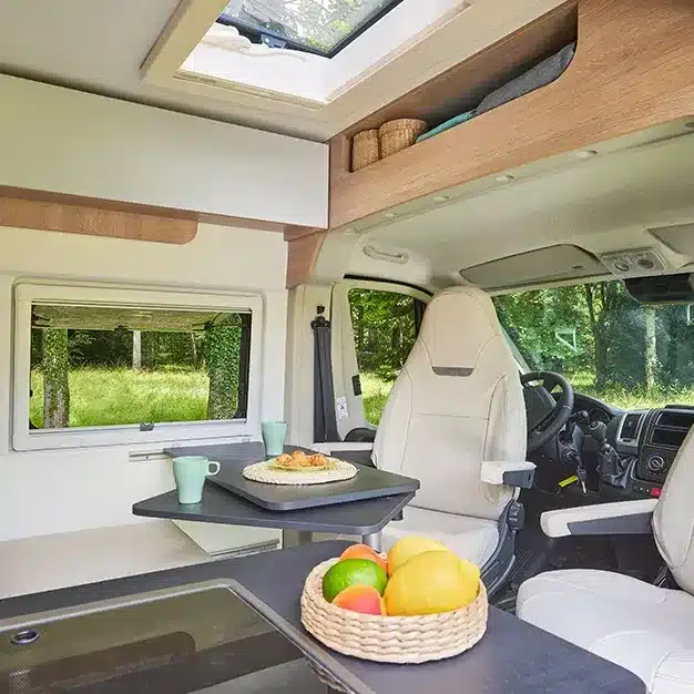 Blick in den hellen Wohnbereich eines Kastenwagens mit Tisch, zwei drehbaren Fahrerhaussitzen und großer Fensterfläche.