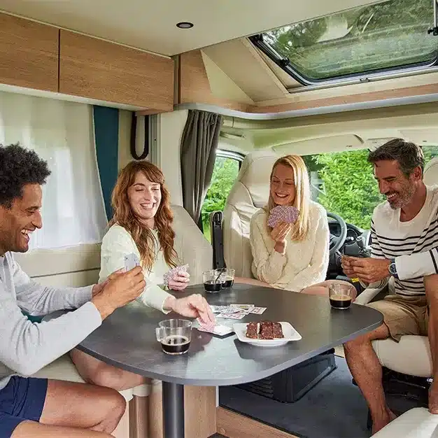 Vier Personen spielen Karten an einem Tisch im Wohnbereich eines Campervans mit viel Tageslicht und modernen Möbeln.