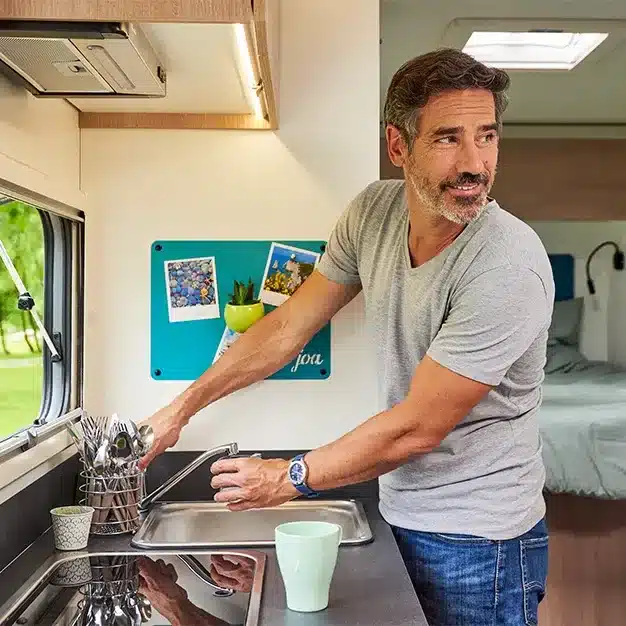 Person spült Geschirr an der Küchenspüle im Camper Joa Camp Teilintegrierte 75T mit grünem Magnetboard und großem Fenster.