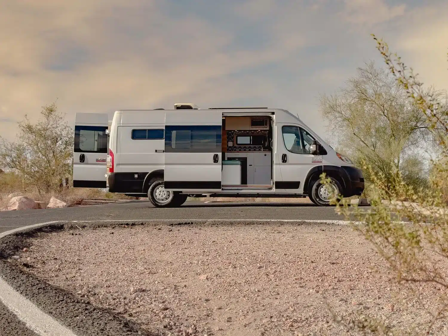 Seitenansicht des Campervans JOLENE mit geöffneten Türen und Einblick in den durchdachten Innenausbau, geparkt in wüstenähnlicher Landschaft.