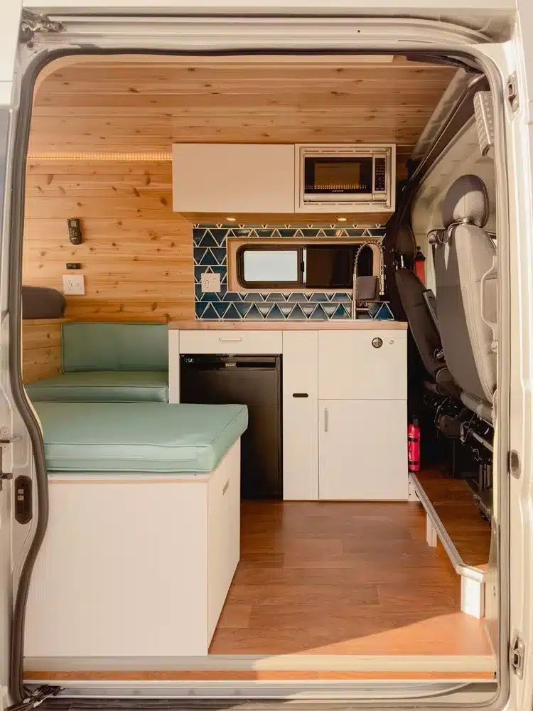 Einblick in das Innere des Campervans JOLENE mit Sitzpolster, Küchenzeile, Kühlschrank und Aufbewahrungsschränken.