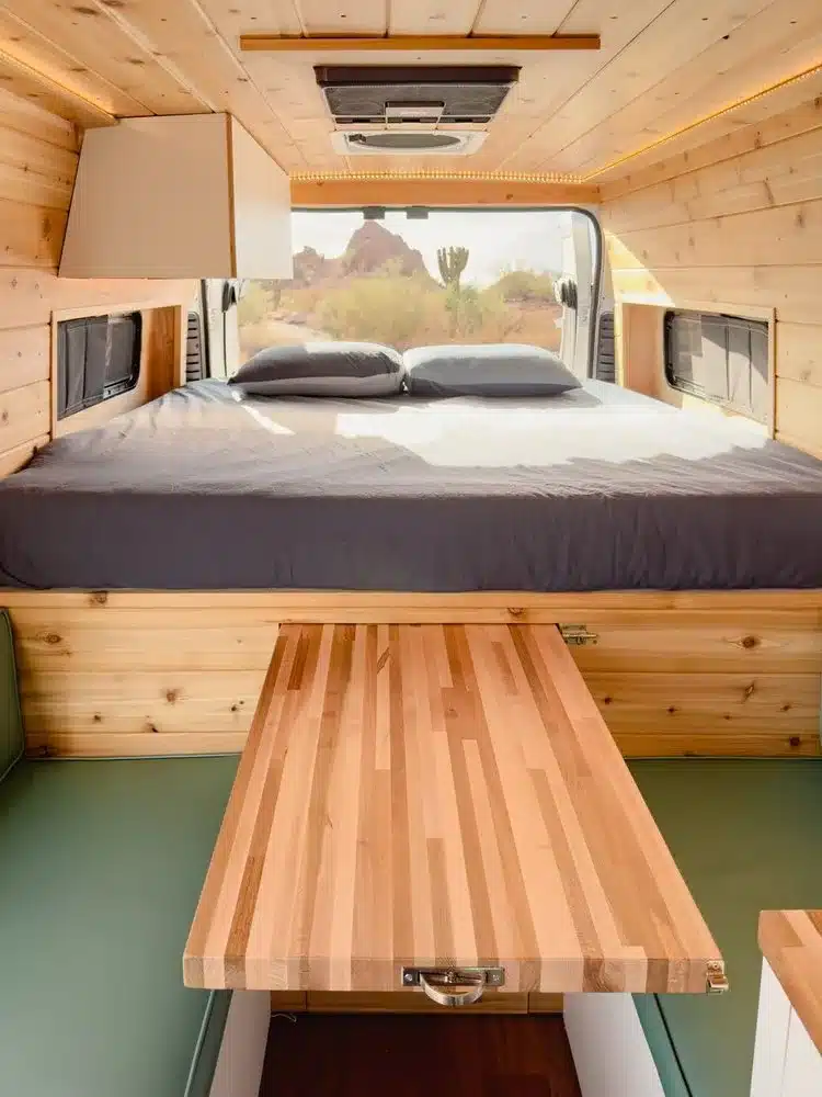 Schlafbereich und ausziehbarer Holztisch im Innenraum des Campervans JOLENE vor offener Hecktür mit Ausblick in die Natur.