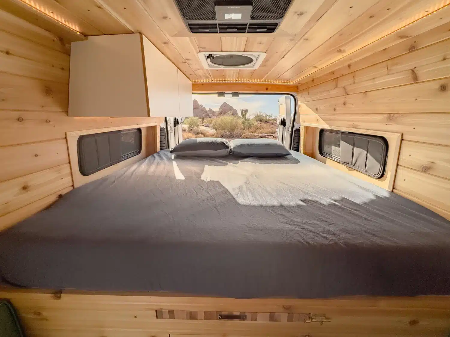 Innenansicht des JOLENE Campervans mit festem Bett, Holzverkleidung und Stauraumfächern.