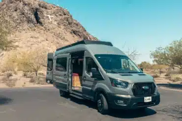 KARUNA Ford Transit Campervan mit geöffneten Türen, geparkt vor felsiger Landschaft unter blauem Himmel.