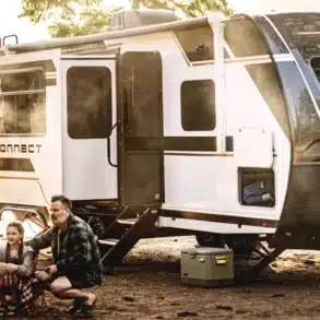 Moderner KZ Connect Travel Trailer beim Camping am Seeufer, umgeben von Bäumen und einer sitzenden Familie am Lagerfeuer.