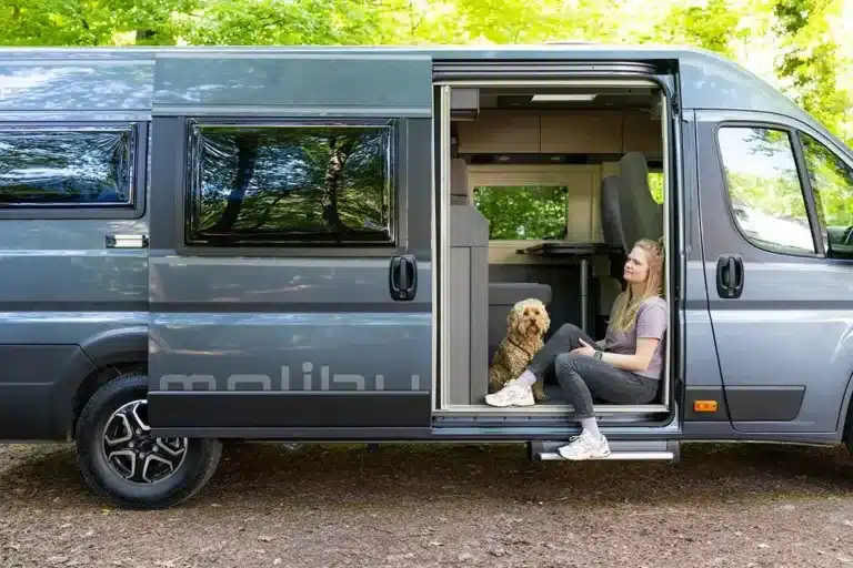 Malibu Relax Kastenwagen in eisengrau, geöffnet, Frau und Hund sitzen am Einstieg im Grünen.