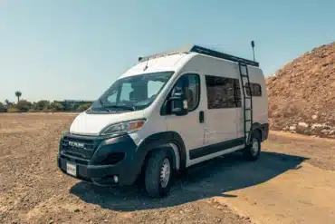 Malvan Ram Promaster 136 High Roof Kastenwagen mit Dachträger und Leiter auf staubigem Untergrund in der Sonne.