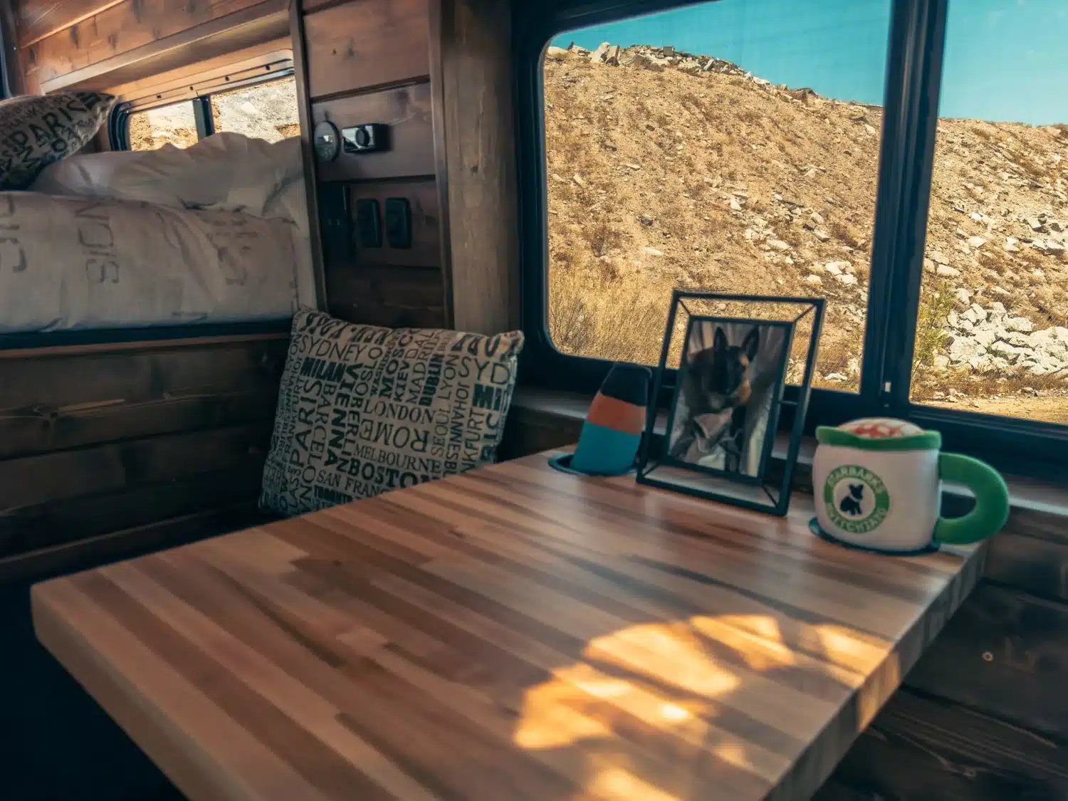 Holztisch und Wohnbereich im Malvan Ram Promaster 136 King Canyon mit Kissen, Tasse und Foto am Fenster