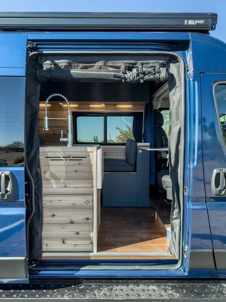 Innenaufnahme des Matt Foley II Campervans mit geöffneter Seitentür, sichtbarer Küchenzeile, Sitzbank und Holzverkleidung.