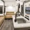Luxuriöser Wohnbereich und Küche im Innenraum der MS Orlando mit weißen Schränken, zwei beigen Sofas, großem Fernseher, Kücheninsel mit Spüle und hochwertigen Geräten.