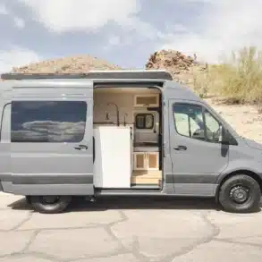 Seitenansicht eines grauen Mercedes-Benz Sprinter 144" mit individuellem Innenausbau und geöffneter Schiebetür in einer trockenen Wüstenlandschaft.