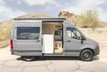 Seitenansicht eines grauen Mercedes-Benz Sprinter 144" mit individuellem Innenausbau und geöffneter Schiebetür in einer trockenen Wüstenlandschaft.