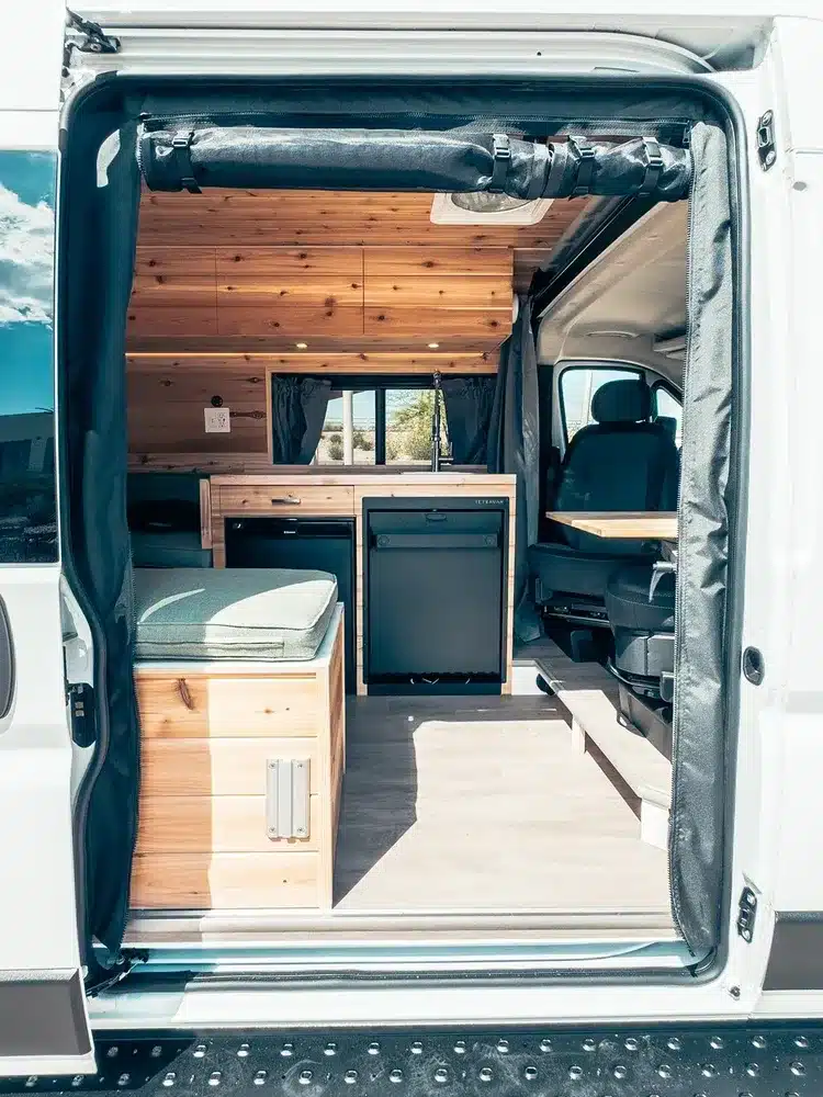 Blick in den Innenraum eines ausgebauten Ram Promaster 2500 Sage Campervans mit Holzausbau, Sitzbank, Küchenzeile und Fenster