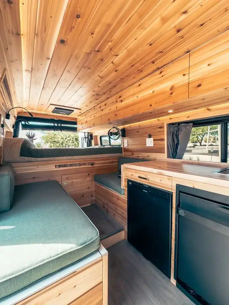 Innenraum eines Sage Campervan mit Holzverkleidung, grünen Polstern, Küchenzeile und Sitzbank