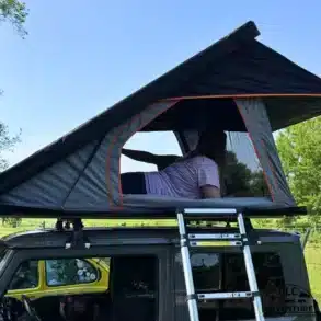 OLC145 Ultra Light Dachzelt mit geöffneter Hartschale und Person darin, montiert auf einem Auto mit Leiter.