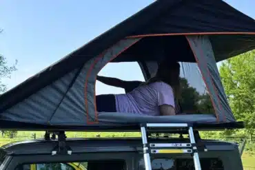 OLC145 Ultra Light Dachzelt mit geöffneter Hartschale und Person darin, montiert auf einem Auto mit Leiter.