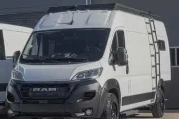 Weißer Ram Falls Campervan mit Dachträger und Außentreppe, geparkt vor einer Halle.