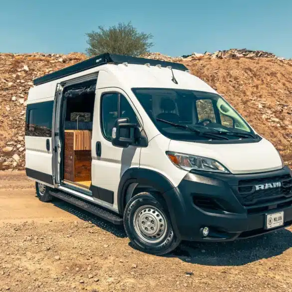 Weißer Ram Promaster 2500 High Roof Campervan mit geöffneter Seitentür, abgestellt auf einer Schotterpiste vor einer steinigen Böschung