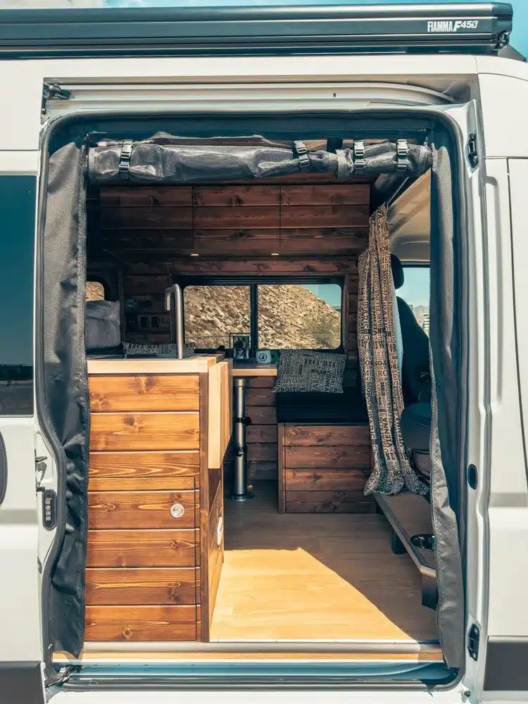 Blick durch die seitliche Schiebetür in den wohnlichen Innenraum eines ausgebauten Ram Promaster 2500 High Roof mit Holzverkleidung und Sitzbereich