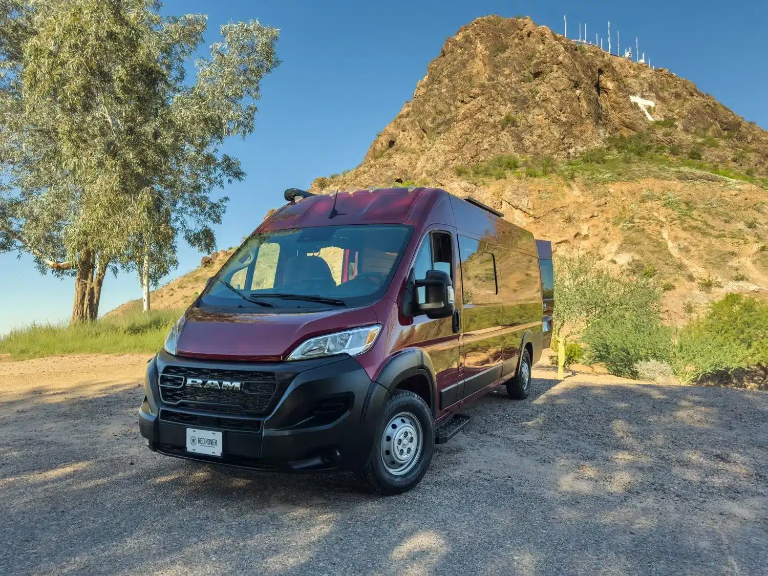 Ein burgunderroter RAM ProMaster 3500 High Roof EXT als Red Rover Campervan steht auf einem Parkplatz vor einem bergigen Hintergrund.