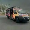 Der Sapho Campervan mit Flammen-Design auf Basis eines RAM Promaster 3500 High Roof, geparkt in wüstenähnlicher Landschaft, Schiebetür geöffnet.