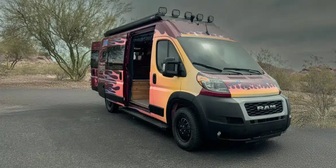 Der Sapho Campervan mit Flammen-Design auf Basis eines RAM Promaster 3500 High Roof, geparkt in wüstenähnlicher Landschaft, Schiebetür geöffnet.