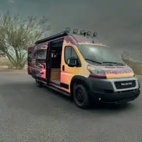Der Sapho Campervan mit Flammen-Design auf Basis eines RAM Promaster 3500 High Roof, geparkt in wüstenähnlicher Landschaft, Schiebetür geöffnet.