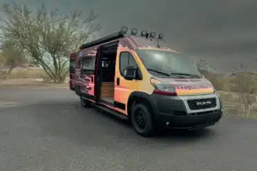 Der Sapho Campervan mit Flammen-Design auf Basis eines RAM Promaster 3500 High Roof, geparkt in wüstenähnlicher Landschaft, Schiebetür geöffnet.