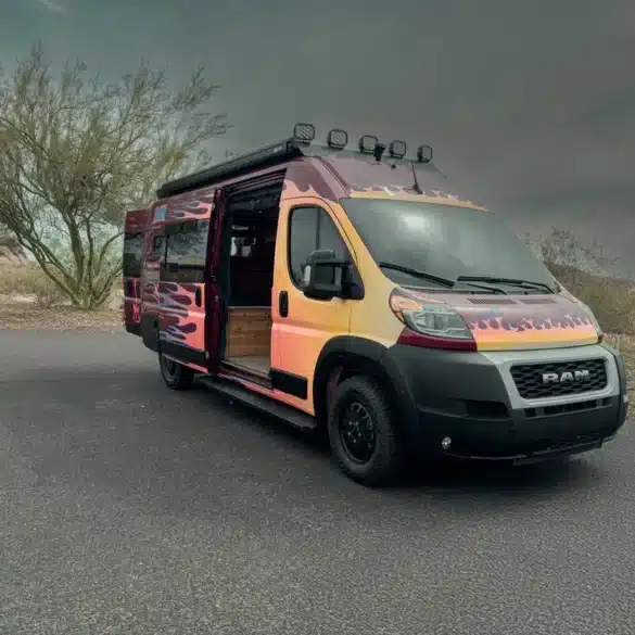 Der Sapho Campervan mit Flammen-Design auf Basis eines RAM Promaster 3500 High Roof, geparkt in wüstenähnlicher Landschaft, Schiebetür geöffnet.