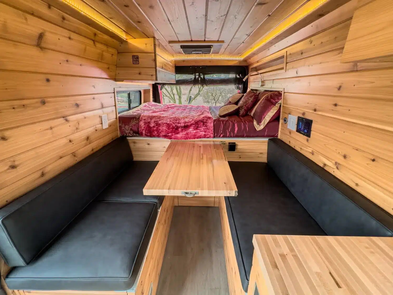 Innenraum eines ausgebauten Kastenwagens mit Holzverkleidung, Sitzgruppe, ausziehbarem Tisch und Bett im Hintergrund