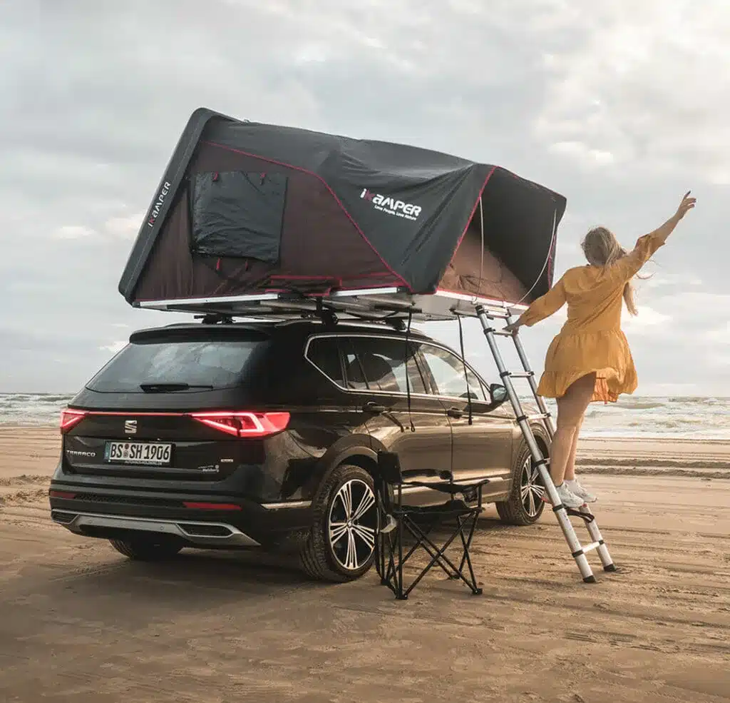 Skycamp 2.0 Dachzelt von iKamper auf einem schwarzen SUV am Strand, geöffnet mit Person auf Leiter.