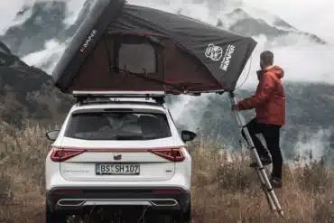 Skycamp 2.0 Dachzelt von iKamper montiert auf einem weißen SUV in einer bergigen Landschaft, eine Person steht auf der Leiter
