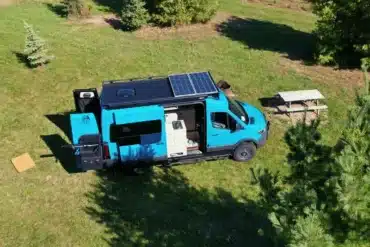 Blauer Sleeping Giant Campervan mit Solarpanels auf dem Dach, von oben fotografiert, auf einer Wiese mit Picknicktisch und Bäumen.