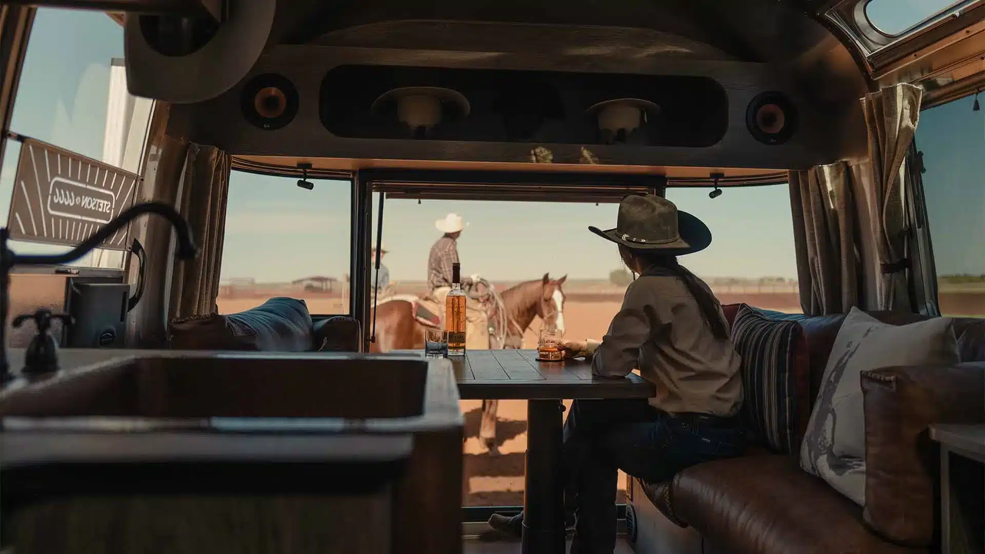 Innenraum eines amerikanischen Travel Trailers mit Western-Motiven; Person mit Hut am Tisch, draußen Cowboys mit Pferden
