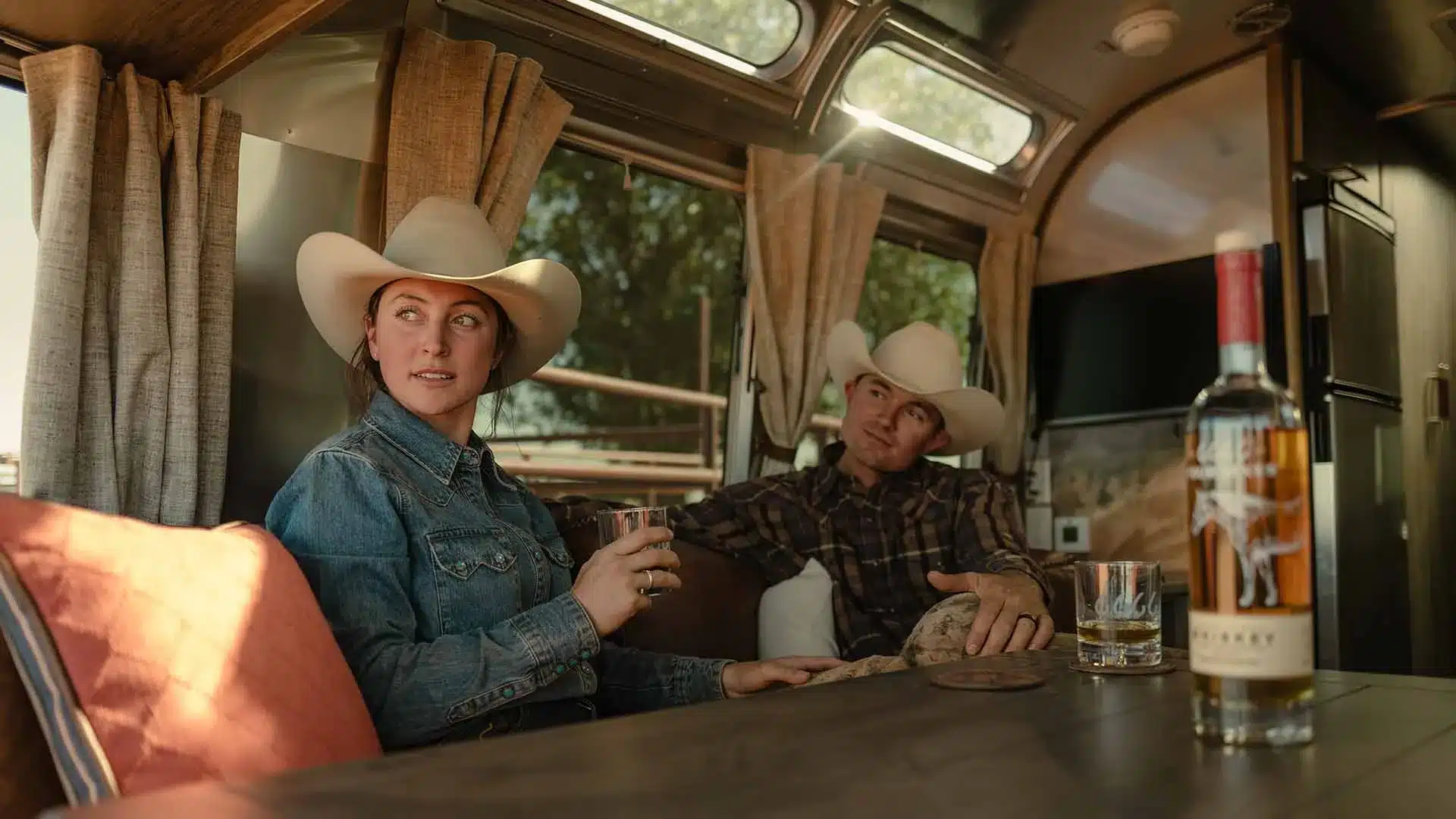 Innenansicht eines amerikanischen Travel Trailers mit zwei Personen im Cowboy-Stil, Sitzbereich mit Western-Details und Getränken auf dem Tisch.