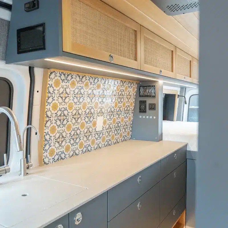 Küchenbereich mit gemusterter Fliesenrückwand und Oberschränken aus Rattan in einem umgebauten Sprinter Kastenwagen.